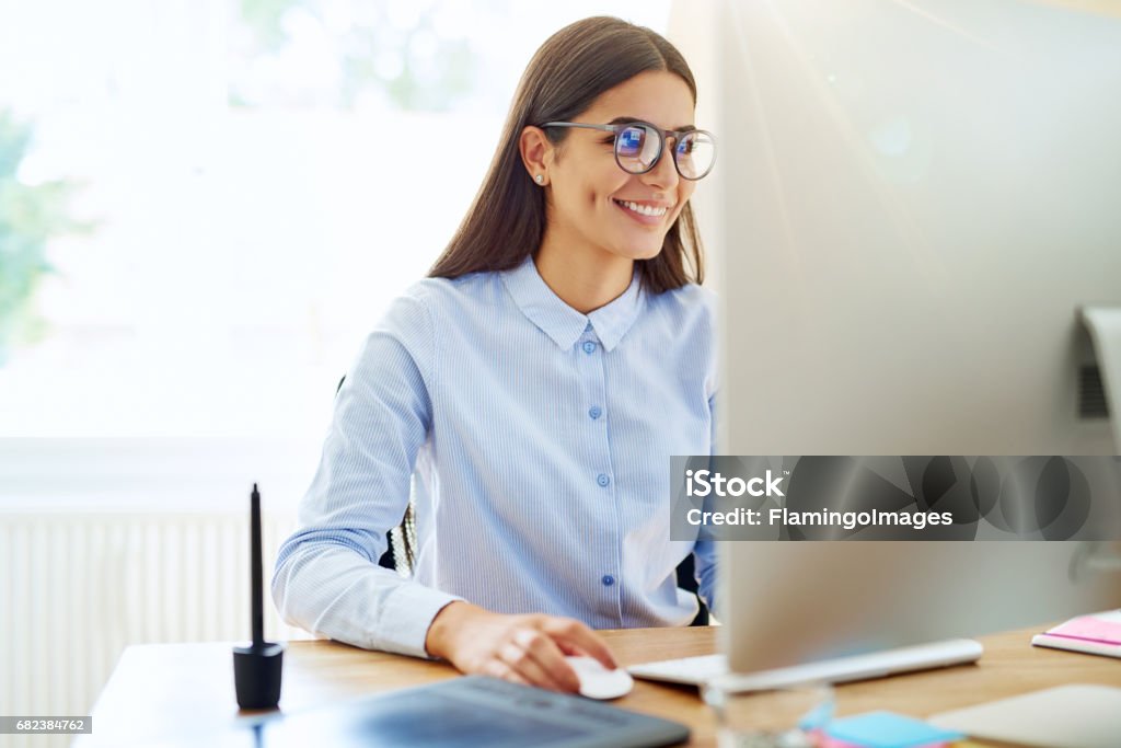 Smiling digital artist working on computer Smiling female graphic designer using mouse, stylus and tablet next to her on wooden desk in small office Desktop PC Stock Photo