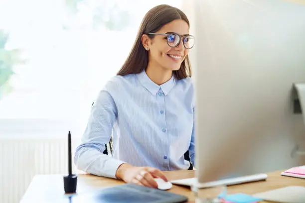 Smiling female graphic designer using mouse, stylus and tablet next to her on wooden desk in small office