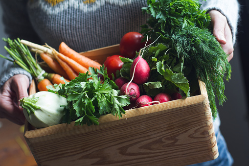 closeup of young Caucasian woman with grey woven sweater holding a large wooden crate full of raw freshly harvested vegetables