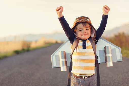 A young male child is standing on a rural Utah road wearing a jet pack, flight cap and flying goggles. He wants to be a pilot when he grows up. He is smiling and is raising his arms. He loves aviation.