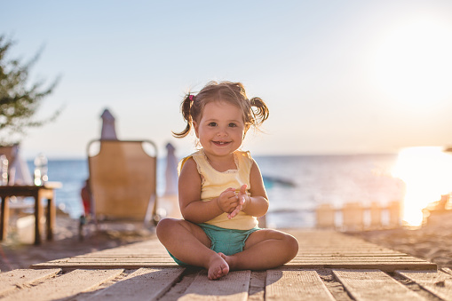 18 month old toddler girl is sitting on the wooden floor of the beach bar. It's her first vacation and she is enjoying the sun and all the fun.