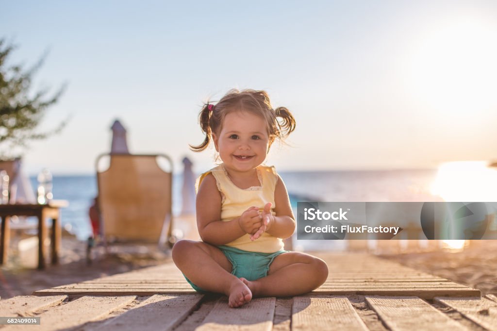 Pretty girl sitting in the sun 18 month old toddler girl is sitting on the wooden floor of the beach bar. It's her first vacation and she is enjoying the sun and all the fun. Baby - Human Age Stock Photo