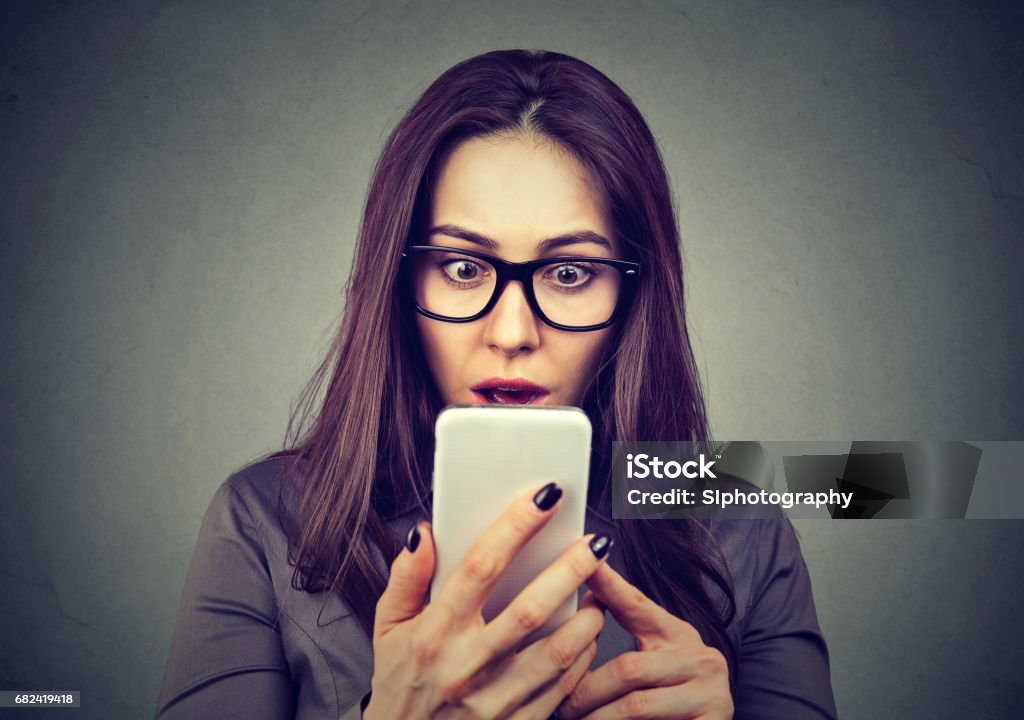 Shocked woman looking at mobile phone Shocked woman looking at mobile phone isolated on gray background Humor Stock Photo