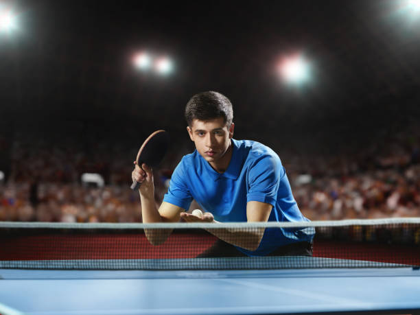 Young ping pong player playing table tennis game. He is holding a red racket. He is very concentrated. Behind him, the stadium with the fans in the defocus.