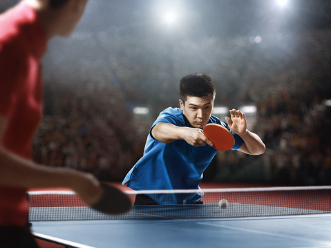 Two ping pong players play table tennis. One is dressed in a red sports uniform, the second is in blue. They are very concentrated.