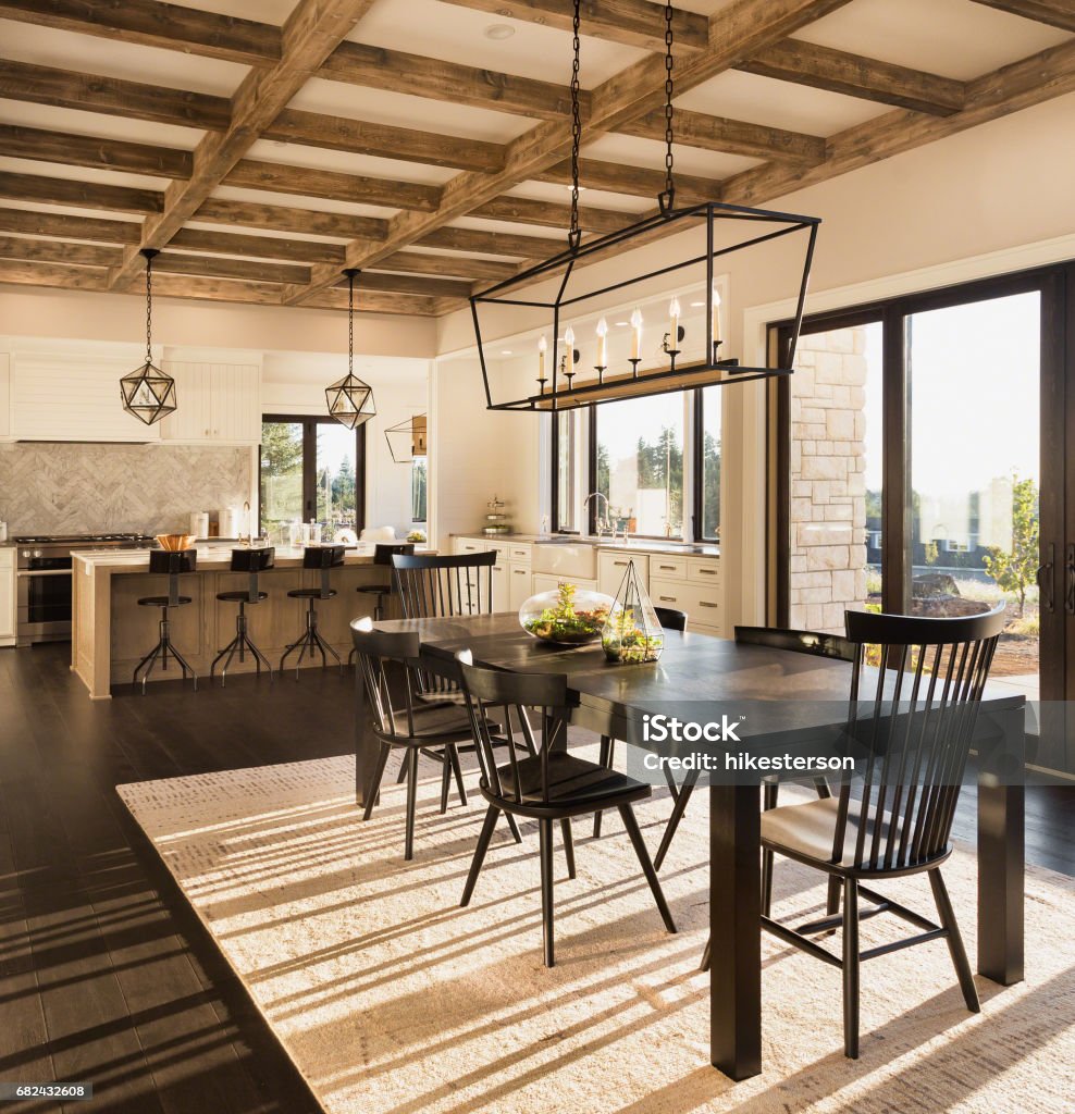 beautiful dining room and kitchen in new luxury home with island and pendant light fixtures Basking in the warmth of a morning sunrise, this luxury dining room and kitchen features elegant pendant light fixtures, glass doors, a wood beam ceiling, and a kitchen island. Is fully furnished Kitchen Stock Photo