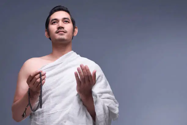 Young asian muslim man wearing ihram clothes with prayer beads against dark background