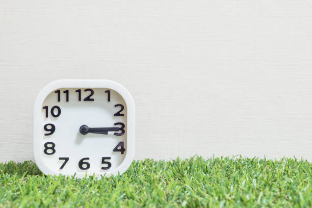 istock Closeup white clock for decorate show a quarter past three o'clock or 3:15 p.m. on green artificial grass floor and cream wallpaper textured background with copy space 682462252