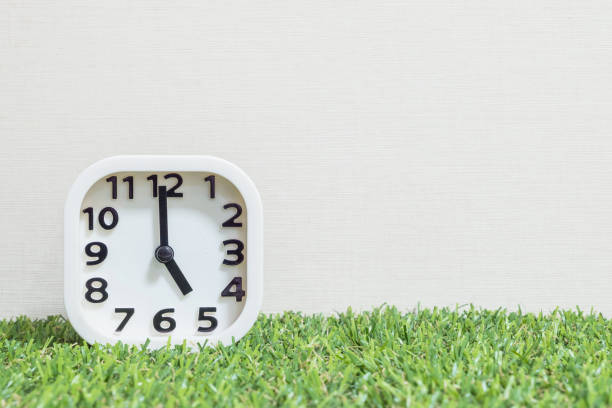istock Closeup white clock for decorate in 5 o'clock on green artificial grass floor and cream wallpaper textured background with copy space 682464440