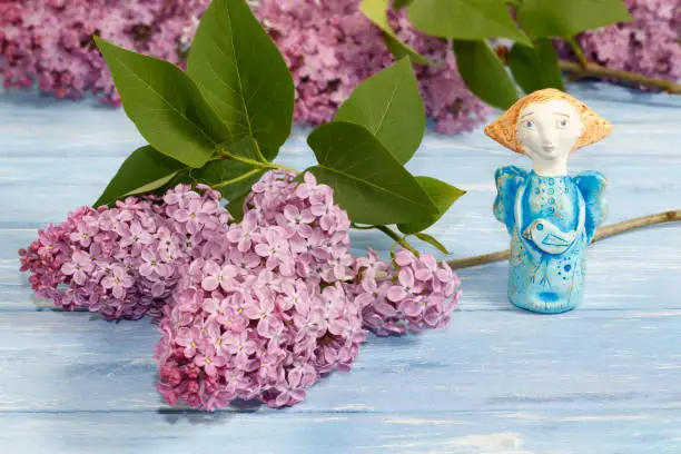 Photo of pottery angel from clay   and branches of lilac on old blue wooden table