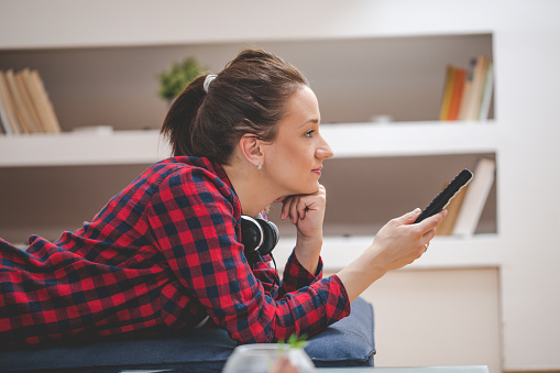 One woman, lying on sofa, listening music on headphones, using remote control.