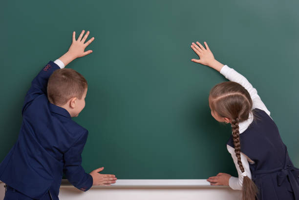 istock elementary school boy and girl put hands on chalkboard background and show blank space, dressed in classic black suit, group pupil, education concept 682518014