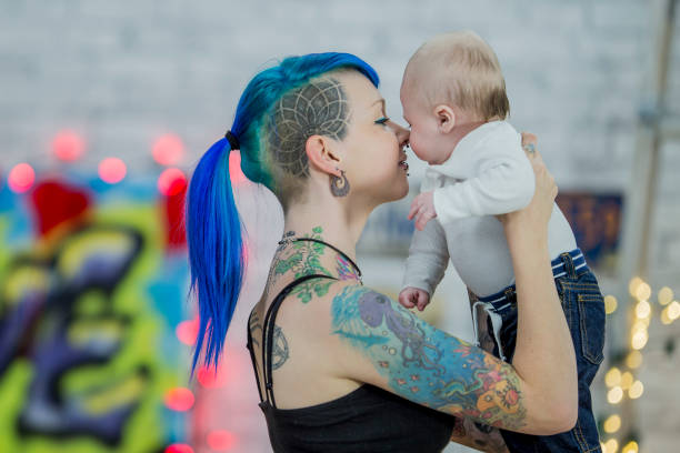 Mother And Son A Caucasian mother and son are indoors. They are in a loft apartment. The mother has blue hair in a punk style, and many tattoos and piercings. She is holding the baby in her arms and putting her nose close to his. alternative lifestyle stock pictures, royalty-free photos & images