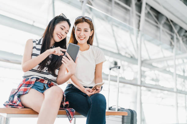 two happy asian girls using smartphone checking flight or online check-in at airport together, with luggage. air travel, summer holiday, or mobile phone application technology concept - japanese girl imagens e fotografias de stock