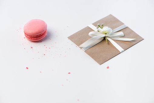 Close-up view of decorative kraft envelope with bow and pink macaron isolated on white, wedding invitation card concept