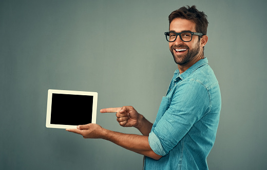 Studio portrait of a handsome young man pointing to a digital tablet with a blank screen against a grey background