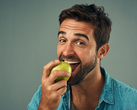 Studio portrait of a handsome young man eating an apple against a grey background