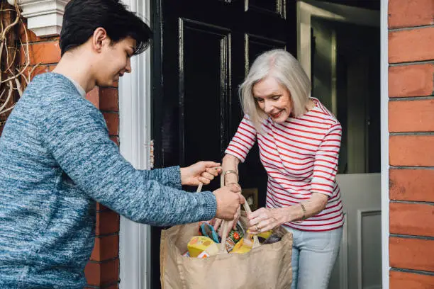 Teenage boy is delivering some groceries to an elderly woman. He is handing her a shopping bag at her front door.