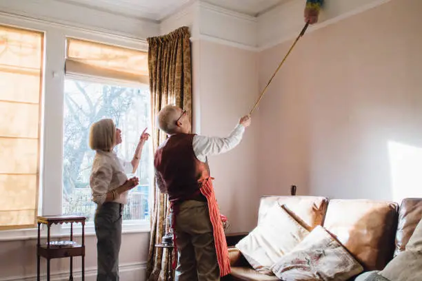 Senior man is helping his wife with the housework. He is using an extended feather duster to clean the ceiling and his wife is pointing out areas he has missed.