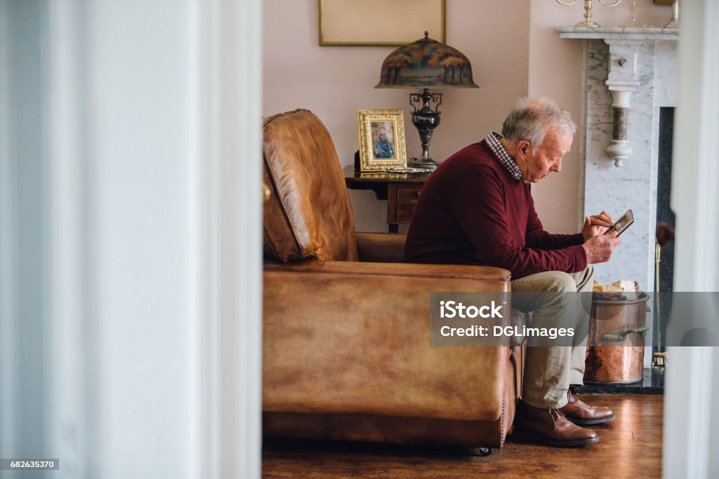 Bringing Back Memories Senior man is sitting in an armchair in the living room of his home, holding and looking at an old photo with a sad expression. Senior Adult Stock Photo