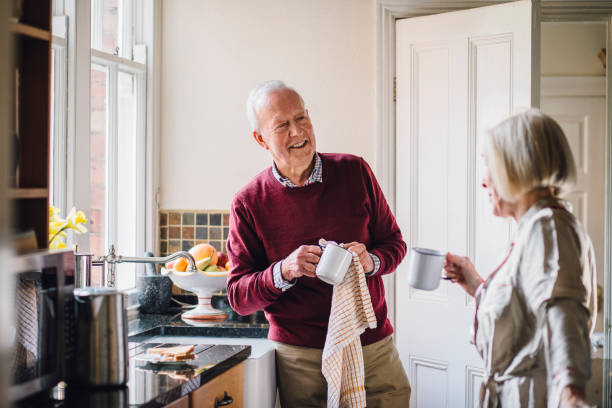 Happy Couple In The Kitchen Senior man is talking to his wife in the kitchen. He is drying dishes and the woman is dirnking tea. senior home stock pictures, royalty-free photos & images