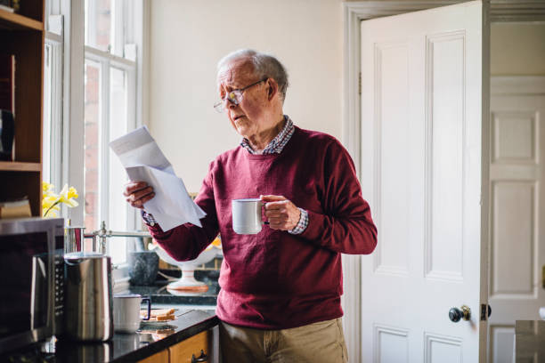 Struggling With Bills Senior man is standing in the kitchen of his home with bills in one hand and a cup of tea in the other. He has a worried expression on his face. cost of living stock pictures, royalty-free photos & images