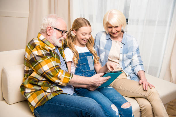 istock Happy grandparents and smiling granddaughter sitting on sofa and using digital tablet 682640608