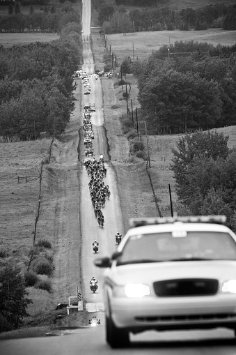 They are lead by a police car and are accompanied by a caravan of race officials, team vehicles with spare bikes and media motorcycles.