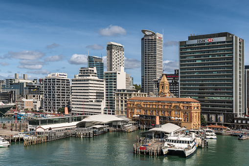 Auckland, New Zealand - March 6, 2017: Ferry building in front of HSBC office building seen from greenish water with a few more high rises in background under blue sky. Ferries at docks.