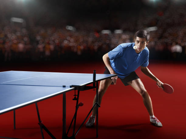 Young ping pong player playing table tennis game. He is holding a red racket. He is very concentrated. Behind him, the stadium with the fans in the defocus.