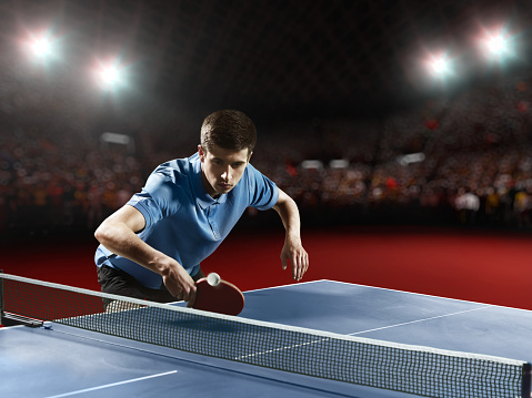 Young ping pong player playing table tennis game. He is holding a red racket. He is very concentrated. Behind him, the stadium with the fans in the defocus.