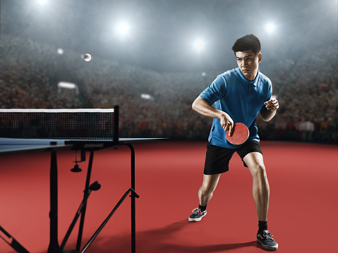 Young asian ping pong player playing table tennis game. He is holding a red racket. He is very concentrated. Behind him, the stadium with the fans in the defocus.
