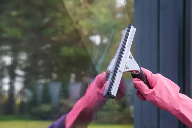 Hand in pink rubber protective glove washing and cleaning window with professionally squeegee. Early spring windows cleaning on the trees background.