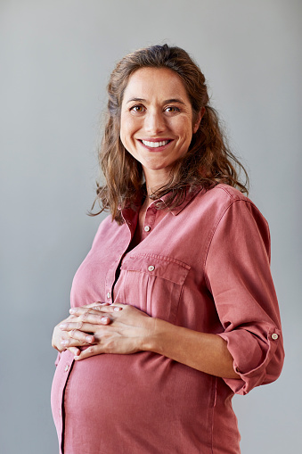 Portrait of pregnant businesswoman with hands on stomach. Female professional is smiling. She is wearing shirt in office.