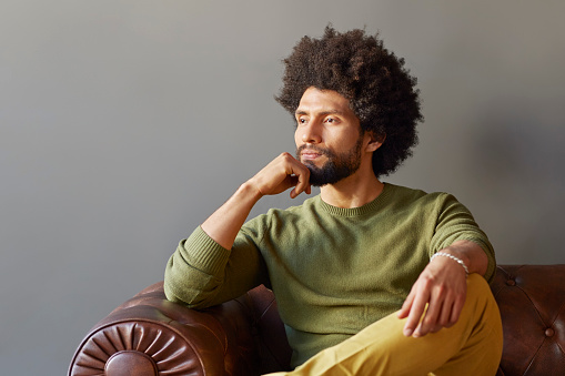 Thoughtful businessman with hand on chin looking away. Male professional is sitting on sofa. He is wearing casuals in office.