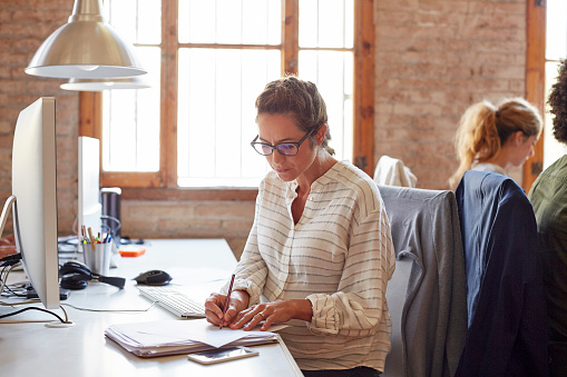 Pregnant woman writing notes at desk. Female executive is sitting on chair. She is working in brightly lit office.