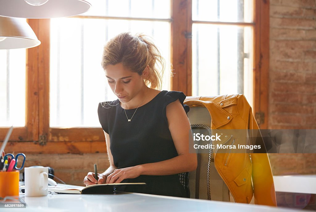 Businesswoman writing in book at desk Young businesswoman writing in book at desk. Female professional is sitting on chair. She is making notes in office. Women Stock Photo