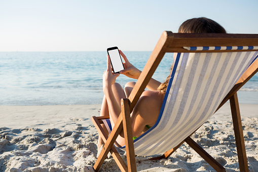 Woman using mobile phone while relaxing on lounge chair at beach during sunny day