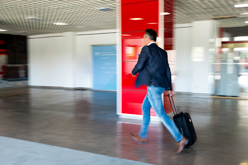 Businessman in a smart casual clothes running with a suitcase in a lobby of a building with a red column. Blurred motion of his moving.