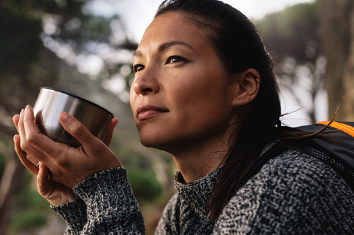 Close up side shot of female hiker resting outdoors with a coffee and looking away. Asian woman having coffee.