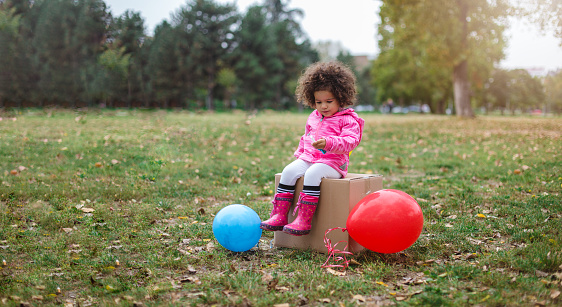Pretty little curly girl is sitting on the cardboard box in the middle on the field. There are two balloons around her, as she is expecting other children to come to an upcoming party.