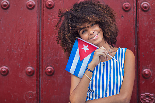 Portrait of happy female holding Cuban flag. Beautiful woman is standing against maroon wooden door. She is wearing striped dress.