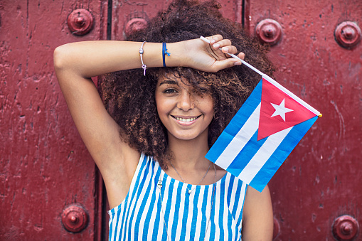 Portrait of happy woman holding Cuban flag. Female is standing against maroon wooden door. She is wearing blue dress.