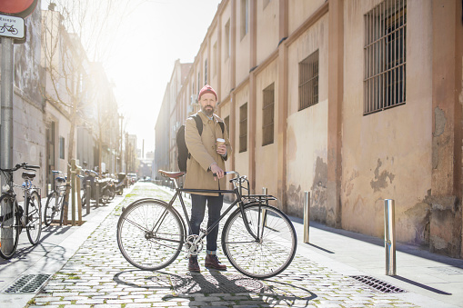 Portrait of man with coffee cup and bicycle. Hipster is standing in city on sunny day. He is wearing jacket.