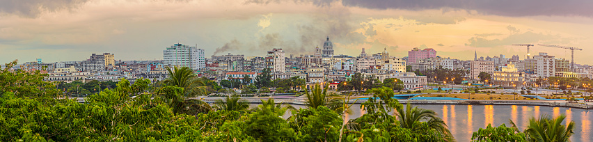 Panoramic view of Havana city against sky. Distant view of Capitolio amidst cityscape during sunset. Buildings and trees are by sea.