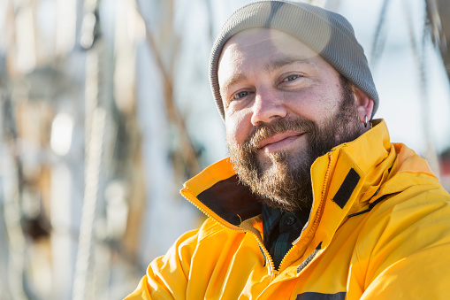 A shrimp boat captain, standing on the deck of his vessel, arms crossed, smiling at the camera wearing a bright yellow raincoat.