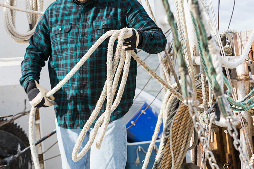 Cropped view of a man wearing a plaid shirt, standing on the deck of a commercial fishing boat, gathering rope. He is unrecognizable with only his midsection showing.