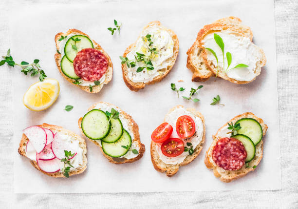 variety of mini sandwiches with cream cheese, vegetables and salami. sandwiches with cheese, cucumber, radish, tomatoes, salami, thyme, lemon zest on a light background, top view. flat lay - crostini imagens e fotografias de stock