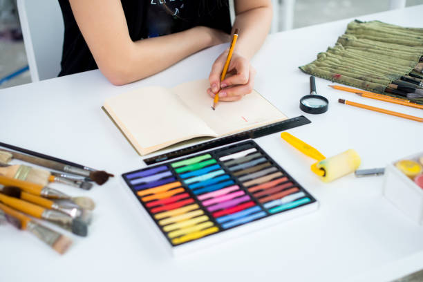 istock Close-up angle view of a female painter drawing draft at sketchbook using pencil. Artist sketching in art studio with crayons set. 683243776