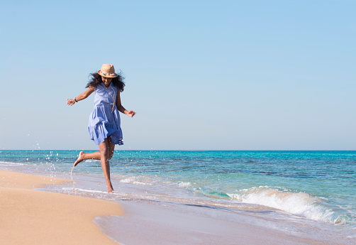 Happy excited woman running, jumping on a sandy sea coast in summer sunny day. Tourist lady wearing blue dress and straw sun hat. Beach holiday concept image.